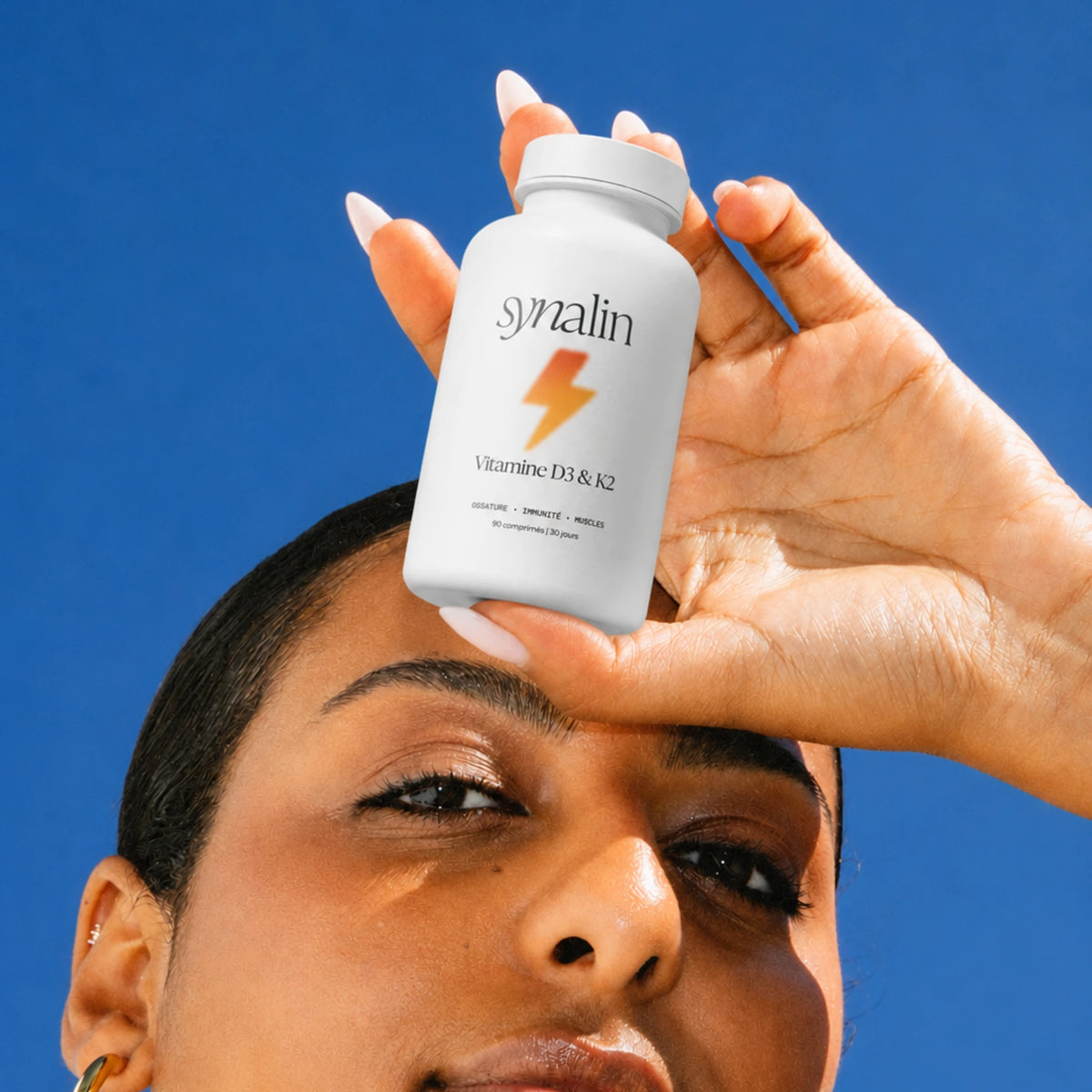 Femme tenant le pilulier Synalin Vitamine D3 & K2 sur fond de ciel bleu